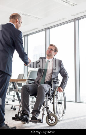 Geschäftsmann Händeschütteln mit lächelnden behinderten Kollegen gegen Fenster im Sitzungssaal an Büro Stockfoto