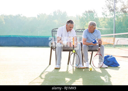 Die volle Länge der Reifen männlichen Freunden sprechen beim Sitzen mit Tennisschläger und Bälle auf Stuhl bei Gericht Stockfoto