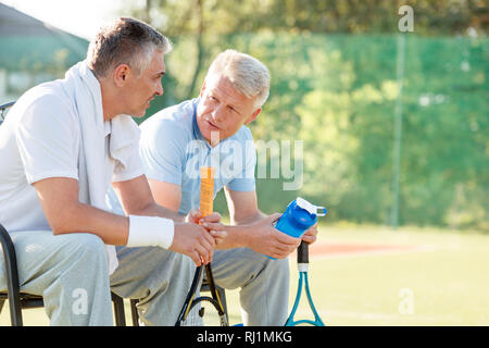 Reife männliche Freunde sprechen beim Sitzen mit Tennisschläger und Wasserflasche auf Stuhl bei Gericht Stockfoto