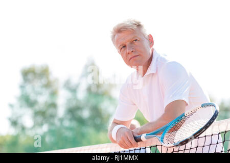 Nachdenklich reifer Mann mit Tennisschläger und lehnte sich auf Net gegen den klaren Himmel Stockfoto