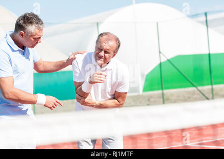 Reifer Mann helfen älteren Freund mit Ellenbogen Verletzung, während Tennis spielen an einem sonnigen Tag Stockfoto