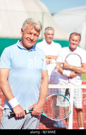 Portrait von zuversichtlich reifer Mann holding Tennisschläger beim Stehen gegen Freunde auf Gericht während der sonnigen Tag Stockfoto