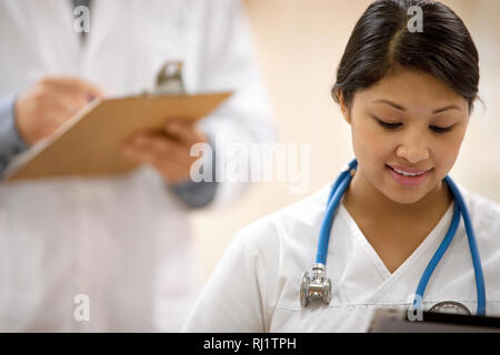 Porträt der jungen weiblichen Krankenschwester Schreiben auf eine Zwischenablage, mit einem männlichen Arzt im Hintergrund. Stockfoto
