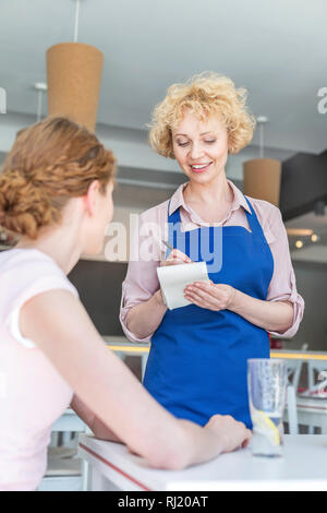 Lächelnde Kellnerin, um von jungen Frau am Tisch sitzen im Restaurant Stockfoto