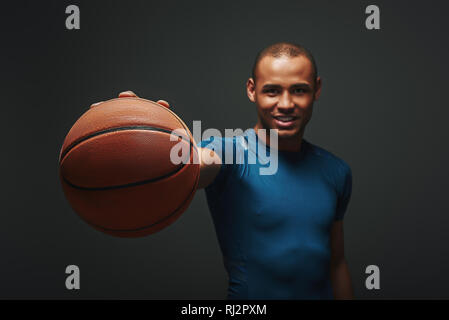 Portrait von stattlichen Basketballspieler in blauen T-Shirt stehend über dunklen Hintergrund isoliert und Kamera. Basketball ist im Fokus Stockfoto
