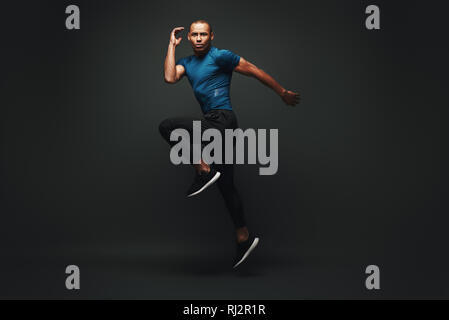 In voller Länge Porträt eines gesunden muskulöse Sportler über dunklen Hintergrund isoliert springen, in die Kamera schaut. Dynamische Bewegung. Seitenansicht Stockfoto