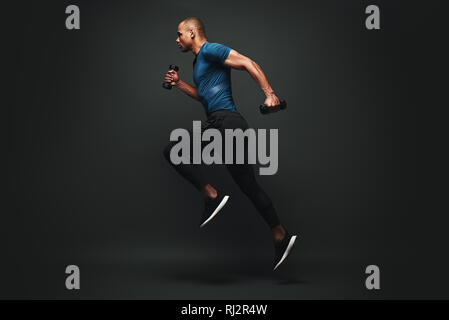 In voller Länge Porträt eines gesunden muskulöse Sportler über dunklen Hintergrund isoliert springen, mit Hanteln in den Händen. Dynamische Bewegung. Seitenansicht Stockfoto