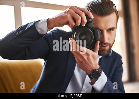 Junge stilvolle Geschäftsmann mit Kamera in den Händen Aufnehmen von Bildern beim Sitzen auf einem Sofa im modernen Büro. Mode suchen. Business Style Stockfoto