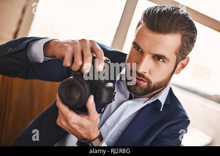 Geschäftsmann mit einer Kamera. Junge Bartgeier und stilvollen Mann ist das Aufnehmen von Fotos mit der Kamera bei einem Besuch im Büro. Business Look. Stockfoto