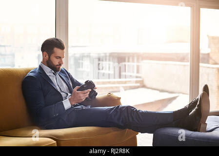 Wohlfühlen... Junge charmante Mann in Mode Anzug ist auf der Suche Fotos von der Kamera und das Gefühl, auf einem Sofa im Büro entspannt. Close-up. Business Look. Stockfoto