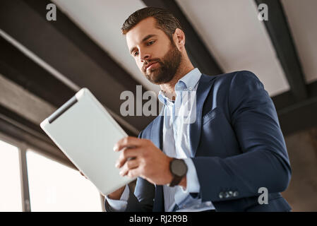 Zeit zu arbeiten! Zuversichtlich bärtige junge Unternehmer im eleganten Anzug wird mit Hilfe von digitalen Tablet-PC im Büro. Business Style. Mode suchen. Stockfoto