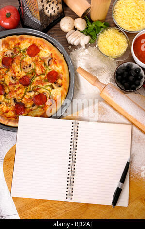 Frisch gebackene Peperoni Pizza mit leeren Notebook oder Kochbuch, hacken, Brett und verschiedenen Zutaten Stockfoto