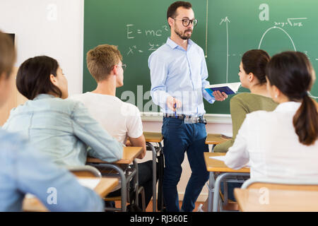 Positiver Mann Lehrer mit Buch gibt interessante Vortrag für Schüler während der Lektion im Klassenzimmer Stockfoto