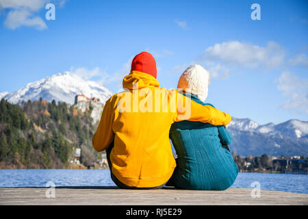 Paar in Liebe umarmen zusammen mit bunten Tüchern sitzen und entspannen auf einer hölzernen Pier auf einem klaren Himmel sonnigen Wintertag Ansicht von der Rückseite, Bled Stockfoto