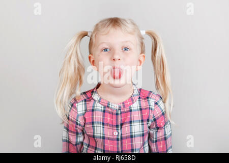 Closeup Portrait von Lustige blonde Kaukasischen Vorschule Mädchen, die Gesichter vor der Kamera. Kind mit Zunge auf normalem hellen Hintergrund. Kid Express Stockfoto