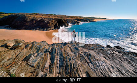 Frau in Wandern Outfit steht auf einer Klippe mit Blick auf einen wilden und natürlichen Strand und Küste mit ruhigem blauen Ozean Stockfoto