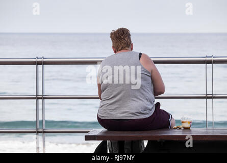 Ansicht der Rückseite des beleibte Frau mit zuckerhaltigen Snack sitzt auf der Bank mit Blick auf das Meer. Stockfoto