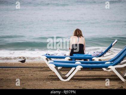 Ansicht der Rückseite Frau mit Zigarette in der Hand sitzen am Strand Liege an einem bewölkten Tag. Stockfoto