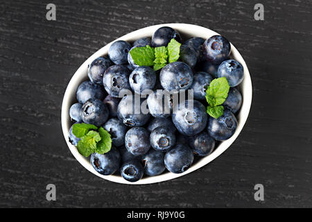 Frische Heidelbeeren in einer weissen Schüssel auf alten schwarzen Holz- Hintergrund, Ansicht von oben (selektive Fokus). Stockfoto