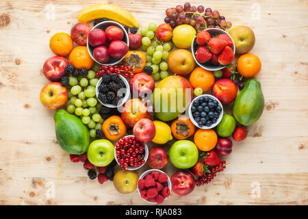 Herz aus gesunden rainbow Früchte, Erdbeeren Himbeeren orangen Pflaumen Äpfel, Kiwis, Weintrauben, Heidelbeeren mango Kaki Ananas auf hellem Holz. Stockfoto