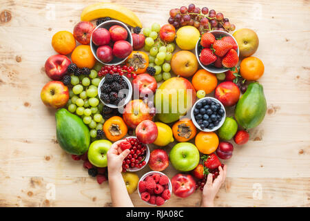 Die Hand des Kindes, die Herzen von gesunden rainbow Früchten, Pflaumen Erdbeeren Himbeeren Orangen, Äpfel, Kiwis, Weintrauben, Heidelbeeren mango Papaya Stockfoto