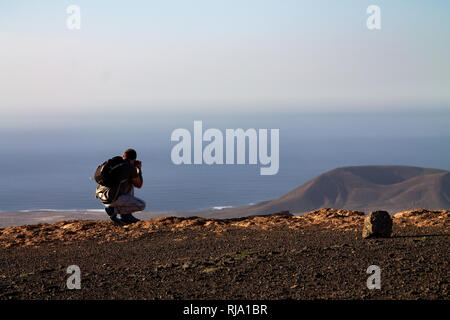 Ein Mann Fotografien auf einer Insel im Ozean, hocken auf dem Rand einer Klippe. Mirador del Rio, Lanzarote, Spanien. Stockfoto