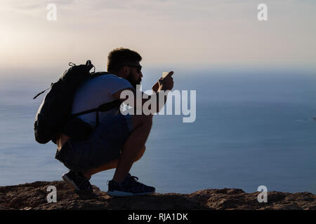 Ein Mann Fotografien auf einer Insel im Ozean, hocken auf dem Rand einer Klippe. Mirador del Rio, Lanzarote, Spanien. Stockfoto