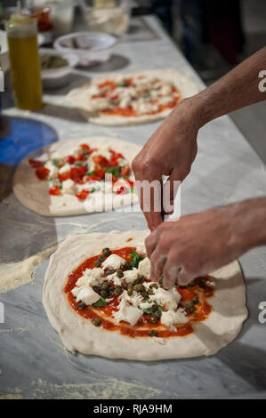 Vorbereitung Pizza Margherita auf einem Ablageflächen aus Marmor. Pizzaiolo setzt Stück Mozzarella, Kapern und Basilikum über eine rohe Pizza Teig. Stockfoto