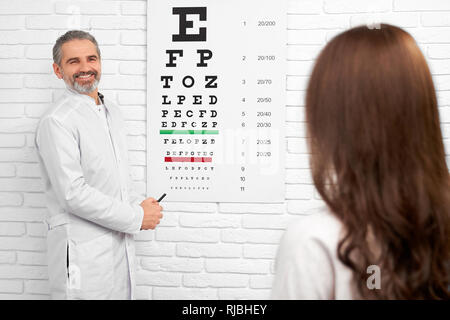 Fröhliche Augenarzt konsultieren die Sehkraft von Patienten in der Klinik. Arzt tragen in Weiß medical Uniform bei Frau suchen, lächelnd, in der Nähe von Eye Test Chart stehen. Stockfoto