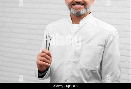 7/8-Portrait von Happy Zahnarzt tragen in Weiß medical Uniform. Bärtige Doktor holding Restaurierung Instrumente. Professionellen Fachmann lächelnd, mit Werkzeugen in den Händen posieren. Stockfoto