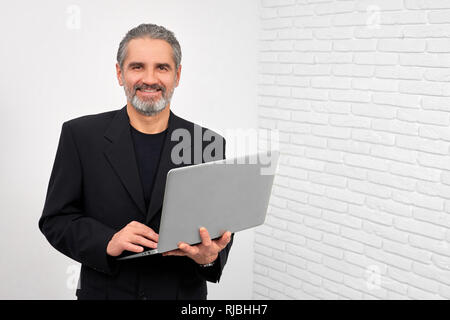 Stattlicher Mann mit grauem Bart und Haare in die Kamera schauen und lächeln. Reifer Mann im Studio mit weißem Hintergrund. Unternehmer halten moderne Notebook, Surfen. Stockfoto