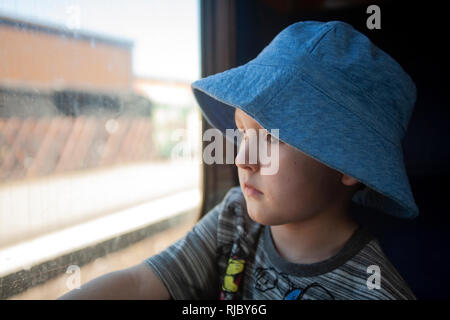 Ein kleiner Junge mit Engelsgesicht, der mit einem blauen Hut verziert ist, fesselt die Reise mit Wunderaugen und blickt aus dem Zugfenster. Stockfoto