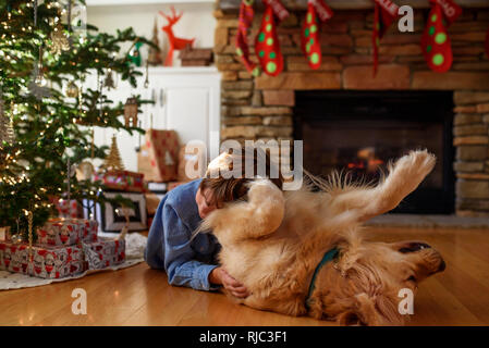 Junge lag auf dem Boden vor einem Weihnachtsbaum seinen Hund kuscheln Stockfoto