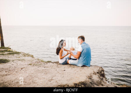 Thema Familienurlaub mit kleinen Kind auf die Natur und das Meer. Mama, Papa und Tochter von einem Jahr sitzen in Umarmung, Mädchen in die Hände des Menschen Stockfoto