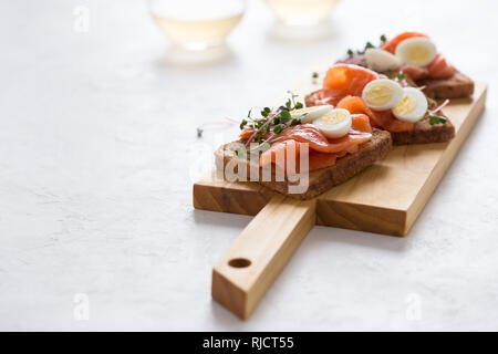 Toast mit geräuchertem Lachs und Wachtelei und Radieschen Sprossen auf rustikalen Holzbrett serviert mit Gläser Weisswein. Köstliche Vorspeise, Snack oder pa Stockfoto