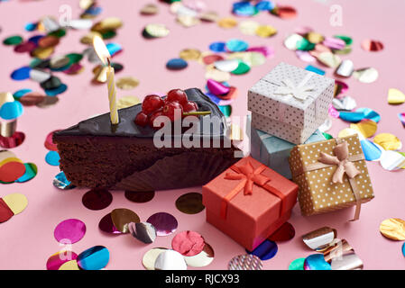 Bereit zum Ausblasen der Kerzen ein Stück Schokoladenkuchen mit Kerze und kleines Geschenk Boxen für ihren Geburtstag. Rosa Hintergrund mit Konfetti Stockfoto