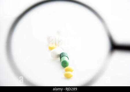 Hand mit Lupe konzentrieren sich auf verschiedene bunte Pillen und Tabletten. Weiß isoliert Hintergrund Stockfoto