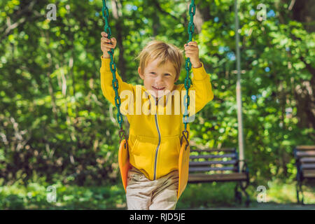 Ein Junge in einem gelben Sweatshirt sitzt auf einer Schaukel auf dem Spielplatz im Herbst. Stockfoto
