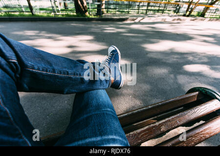 Der Kerl in Jeans und Turnschuhe sitzt auf einer Holzbank im Park. First-Person anzeigen. Stockfoto