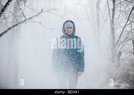 Ein Kerl, der eine Jacke mit Kapuze steht in einem verschneiten Wald, Schnee fällt auf ihn von oben. Stockfoto