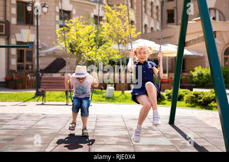 Junge in die Brille und Hut und Blonde Mädchen im Kleid Spaß auf einer Schaukel zusammen in schönen Sommergarten an warmen und sonnigen Tag im Freien. Stockfoto