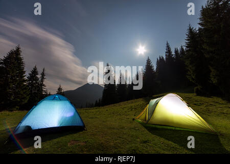 Hellen Mond in Dunkelblau bewölkter Himmel über zwei touristische Zelte auf der grünen Wiese Wald Lichtung unter hohen Kiefern auf ferne Berge im Hintergrund. T Stockfoto