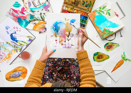 Blick von oben auf die Frau mit Zeichnung auf Knie und Malen mit Wasserfarben malt, während durch farbige Bilder umgeben Stockfoto
