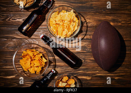 Blick von oben auf die Snacks in Schalen in der Nähe der braunen Flaschen mit Bier und Ball auf hölzernen Tisch Stockfoto