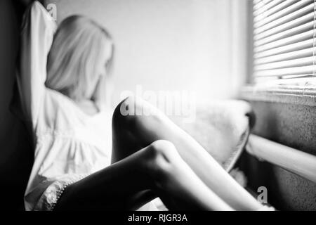 Verschwommene portrait einer Langbeiniges Mädchen sitzen in einem Kleid am Fenster. Ein Buch lesen zu Hause. Stockfoto