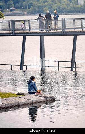 Montreal, Kanada - Juni, 2018: Junge männlich liest ein Buch sitzen am Fluss. Er nahm seine Schuhe und seine Füße im Wasser in Mont Stockfoto