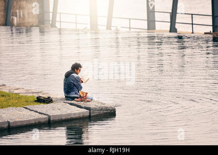 Montreal, Kanada - Juni, 2018: Junge männlich liest ein Buch sitzen am Fluss. Er nahm seine Schuhe und seine Füße im Wasser in Mont Stockfoto