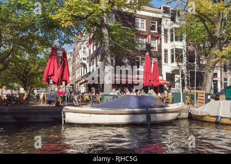 Amsterdam, Niederlande, 10. Oktober 2018: Blick von Canal auf der Terrasse des Cafe mit Menschen ihre Mahlzeiten genießen Sie bei schönem Wetter Stockfoto