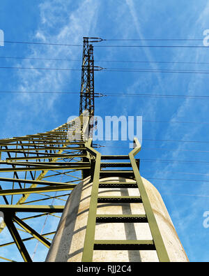 Ansicht von unten bis zu einer hohen Spannung Pylon, der auf einem Betonfundament steht, eine Leiter führt. Über ihm ein blauer Himmel mit Wolken von Nebel. Stockfoto