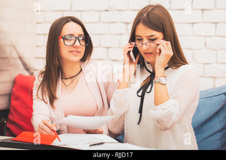 Junge Unternehmerinnen Dokumente gemeinsam diskutieren. Schöne Frau Suche auf dem Papier Dokumente Informationen, während ihr Begleiter spricht über Mobiltelefon Stockfoto